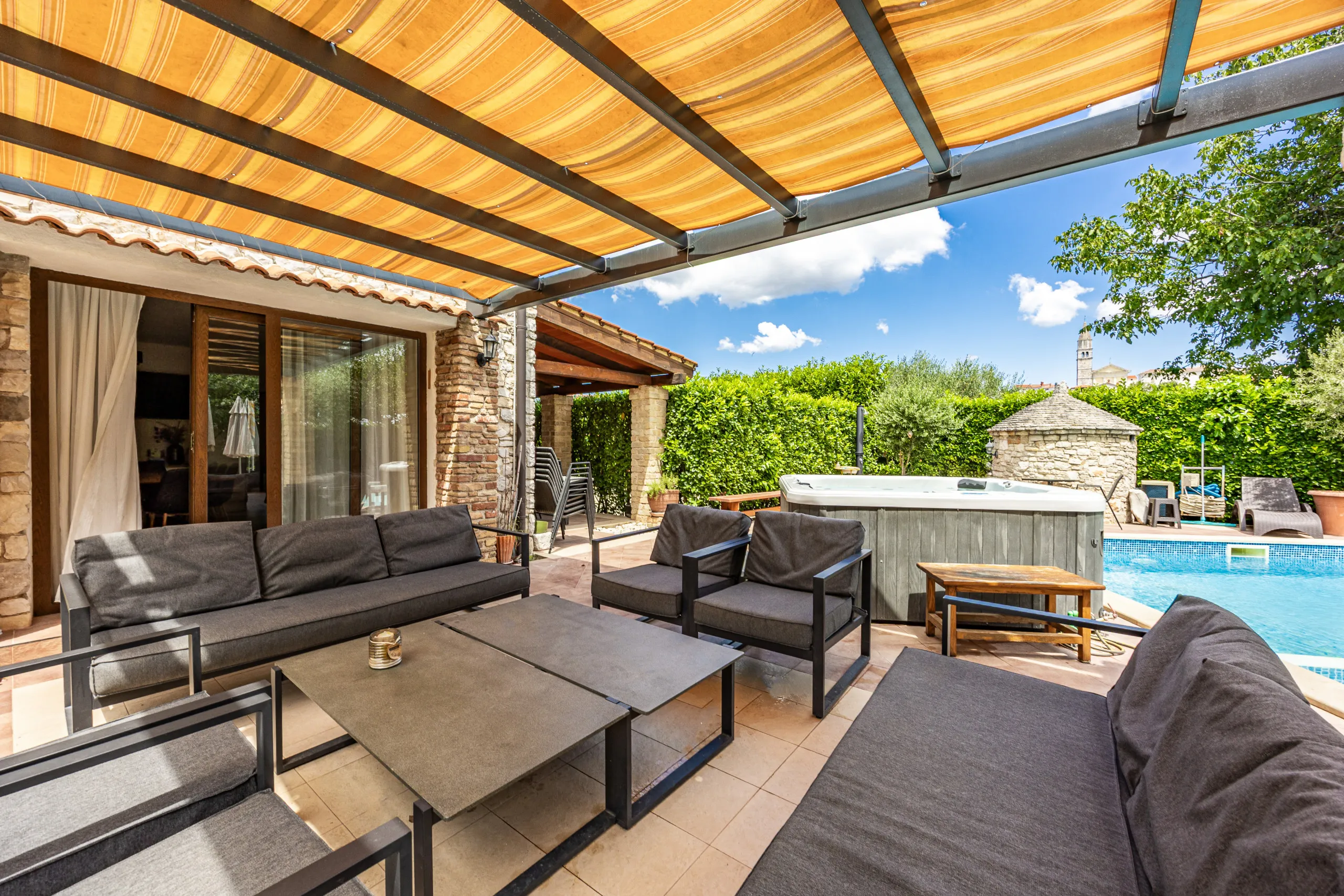 Sofas and low tables under the striped pergola awning, hot tub and pool alongside