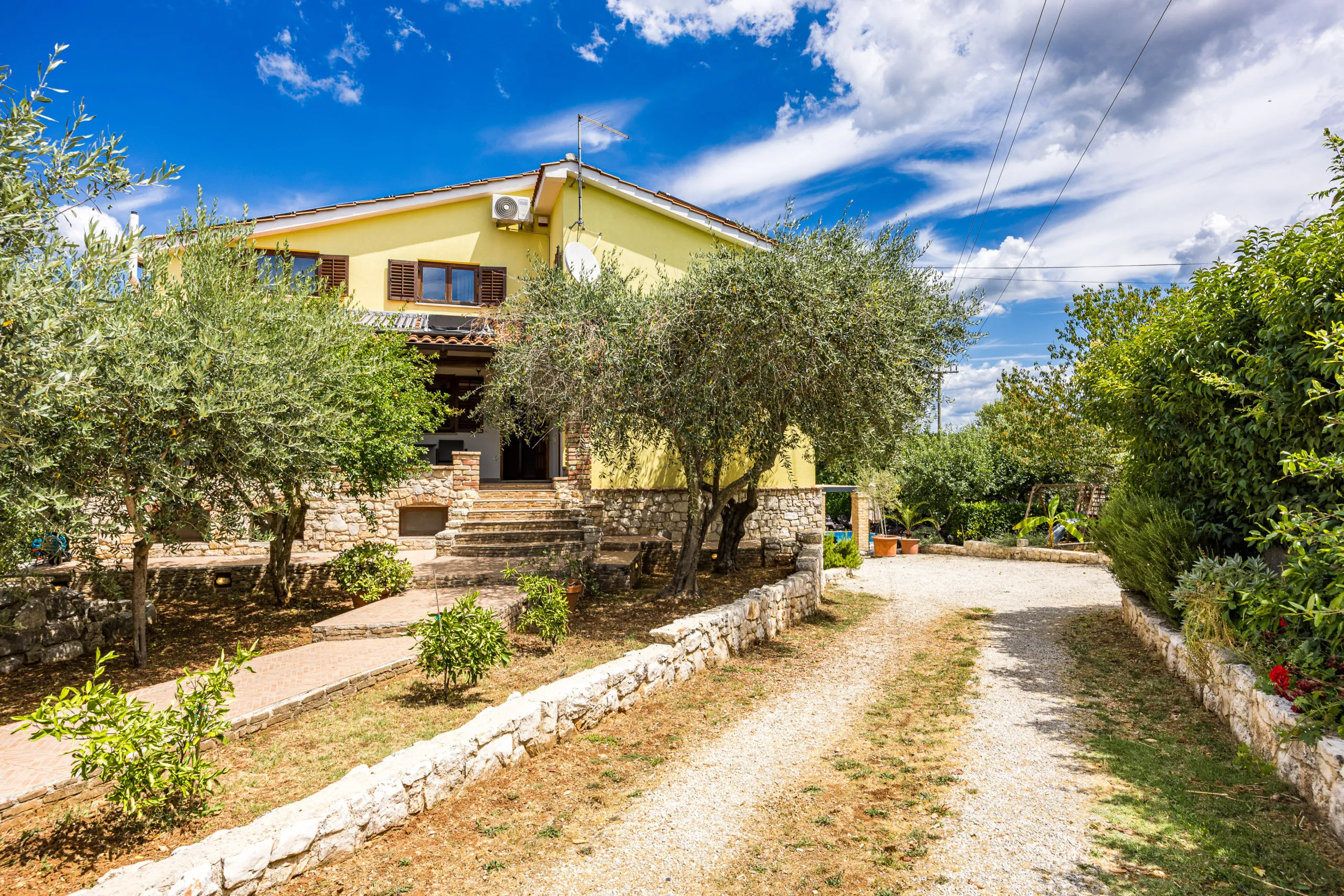 The house above its stone retaining wall, olive trees along the gravel drive