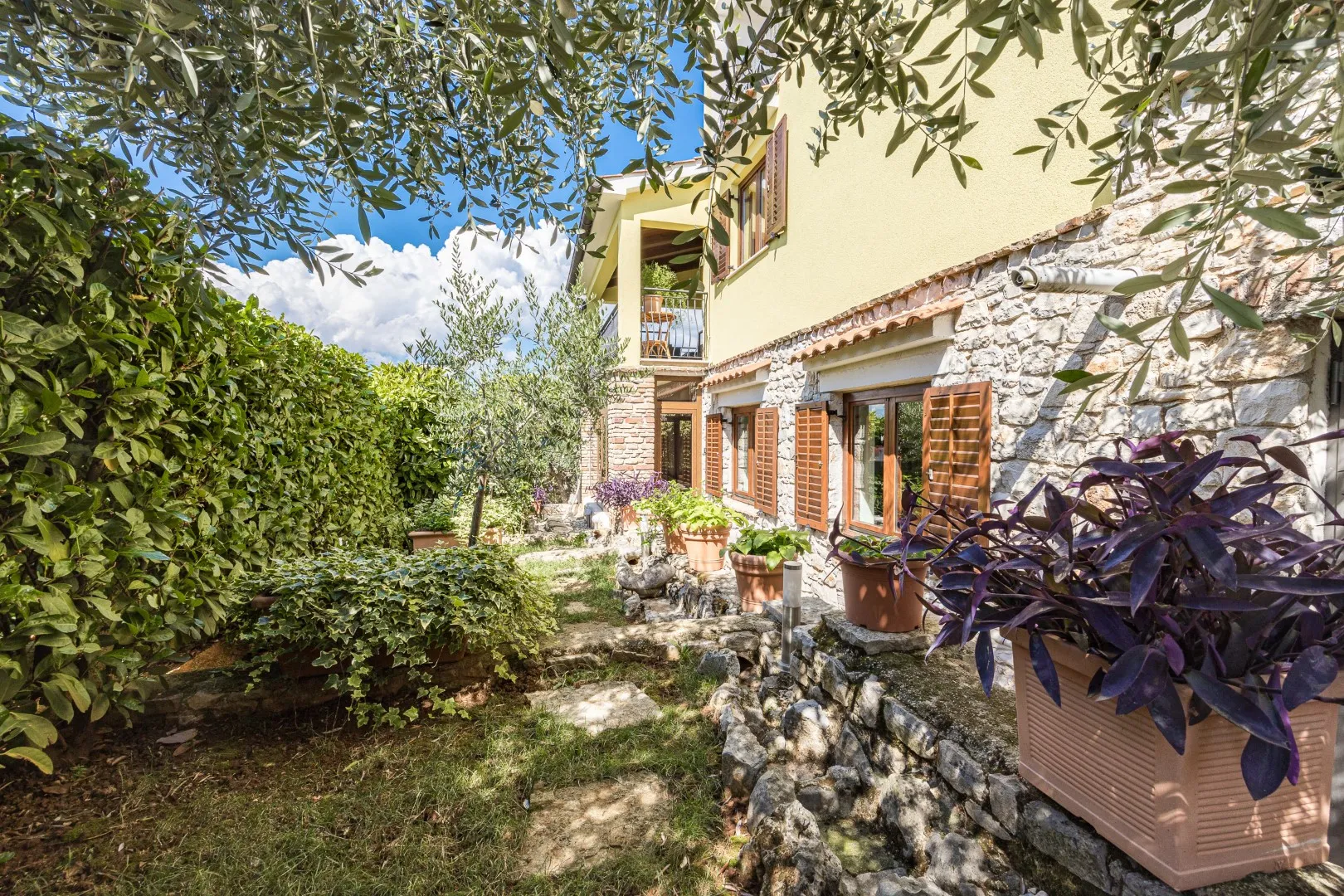 The stone flank of the house, shuttered windows above a planted path