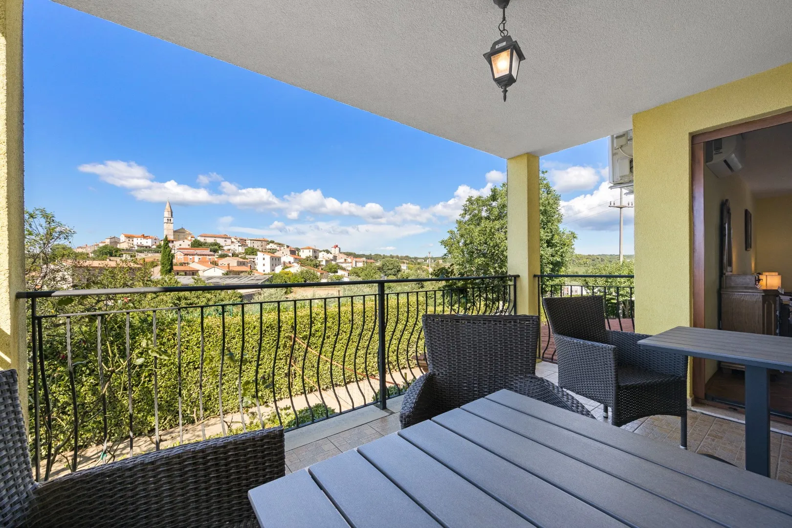 A covered balcony with wicker chairs and a table, looking across Višnjan's rooftops to the church tower