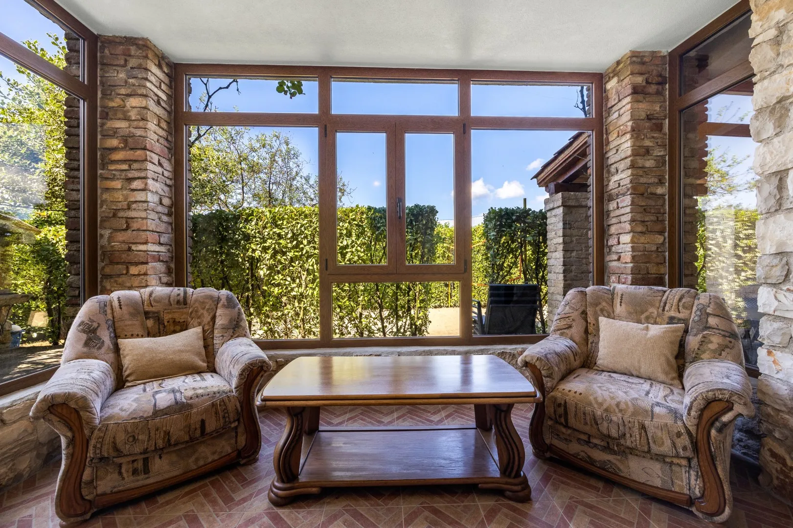 The brick-and-glass sunroom, two armchairs facing the hedge and garden