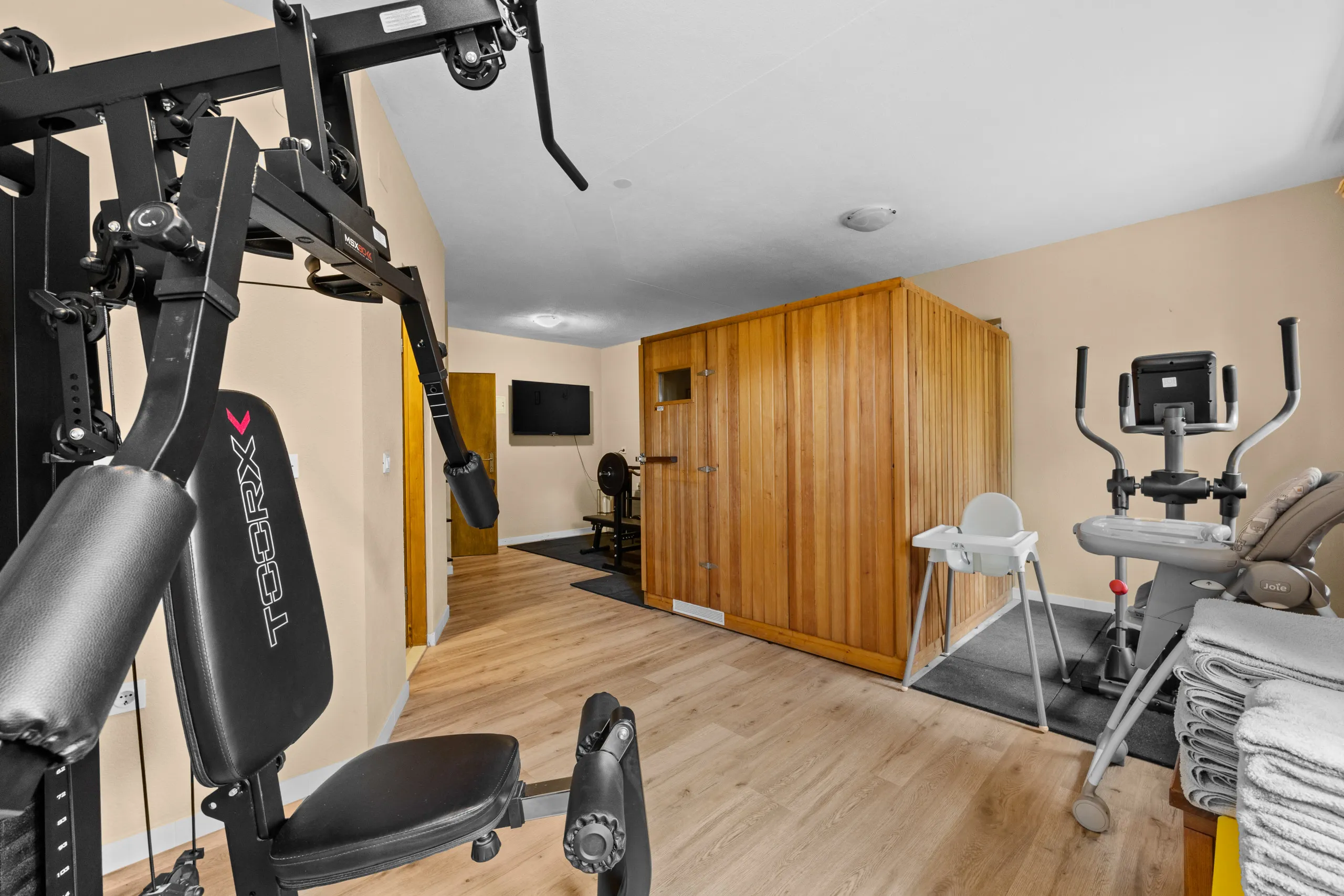 The fitness room with a multi-gym, a cross-trainer and a weights bench, the sauna cabin beside them