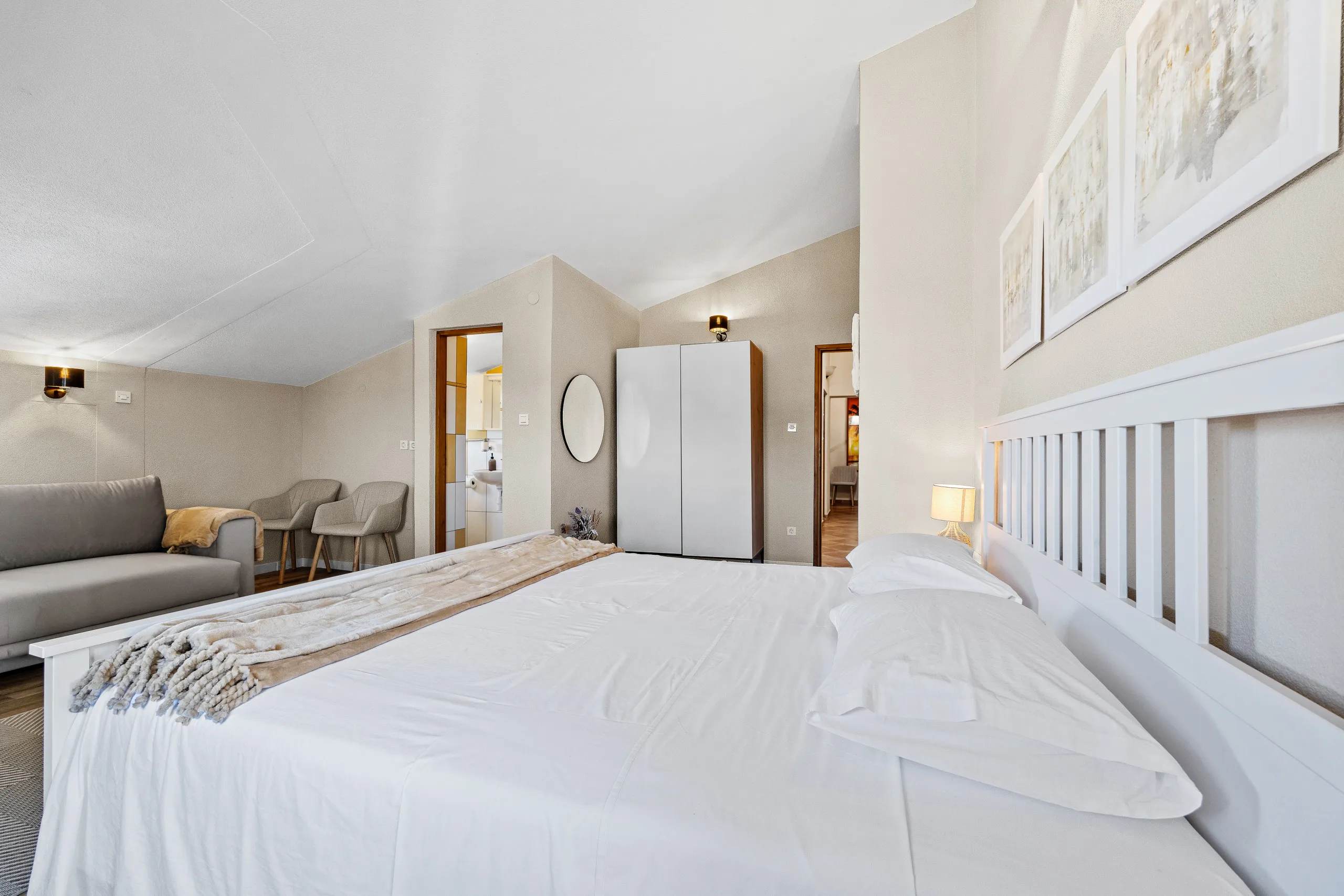 A double bed against a slatted white headboard, sofa and armchairs at the far end