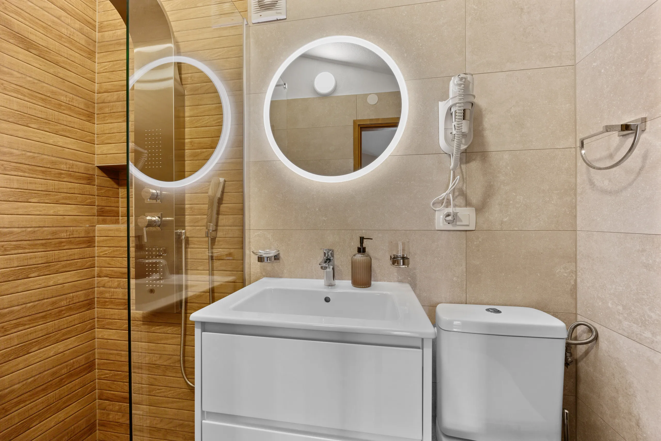 A round backlit mirror above a white vanity, shower cubicle alongside