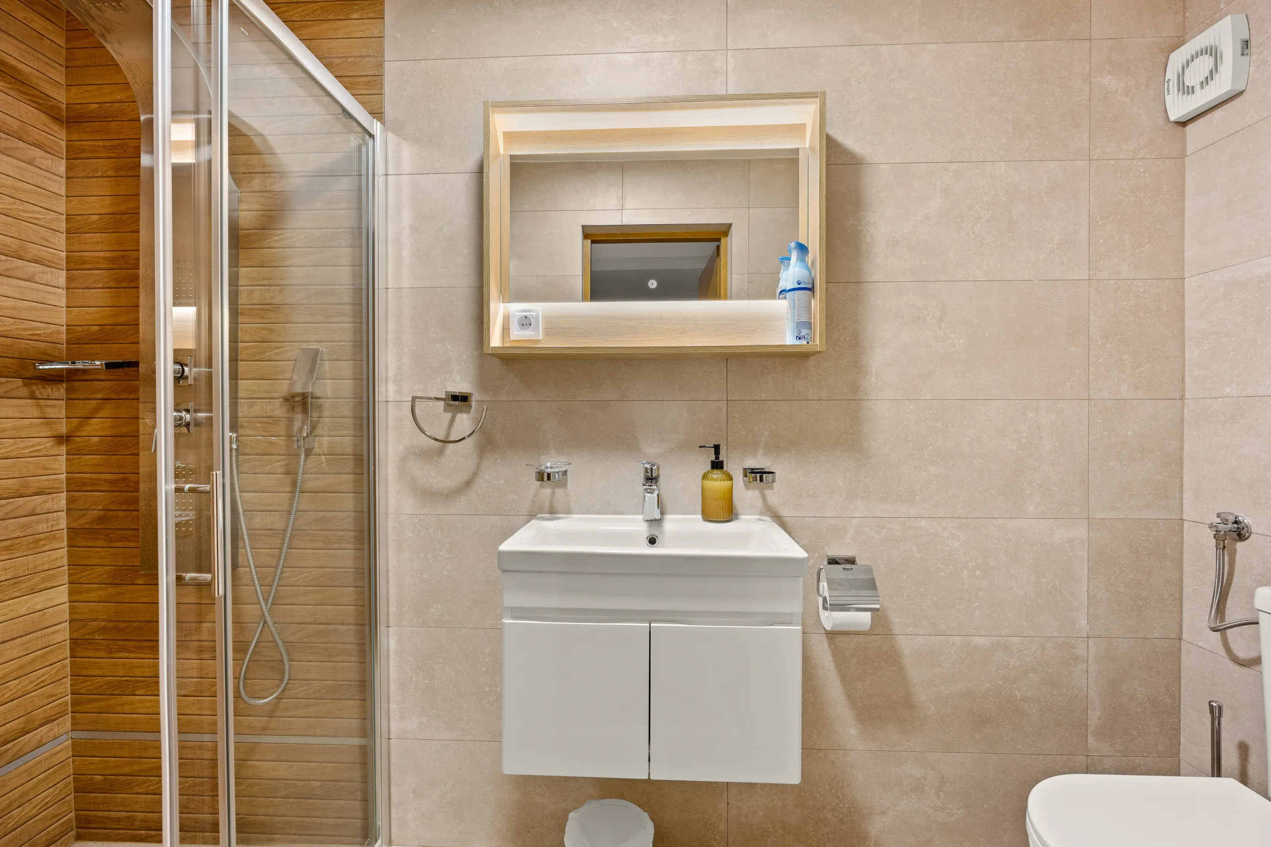 A shower cubicle beside a wall-hung basin and a lit oak-framed mirror