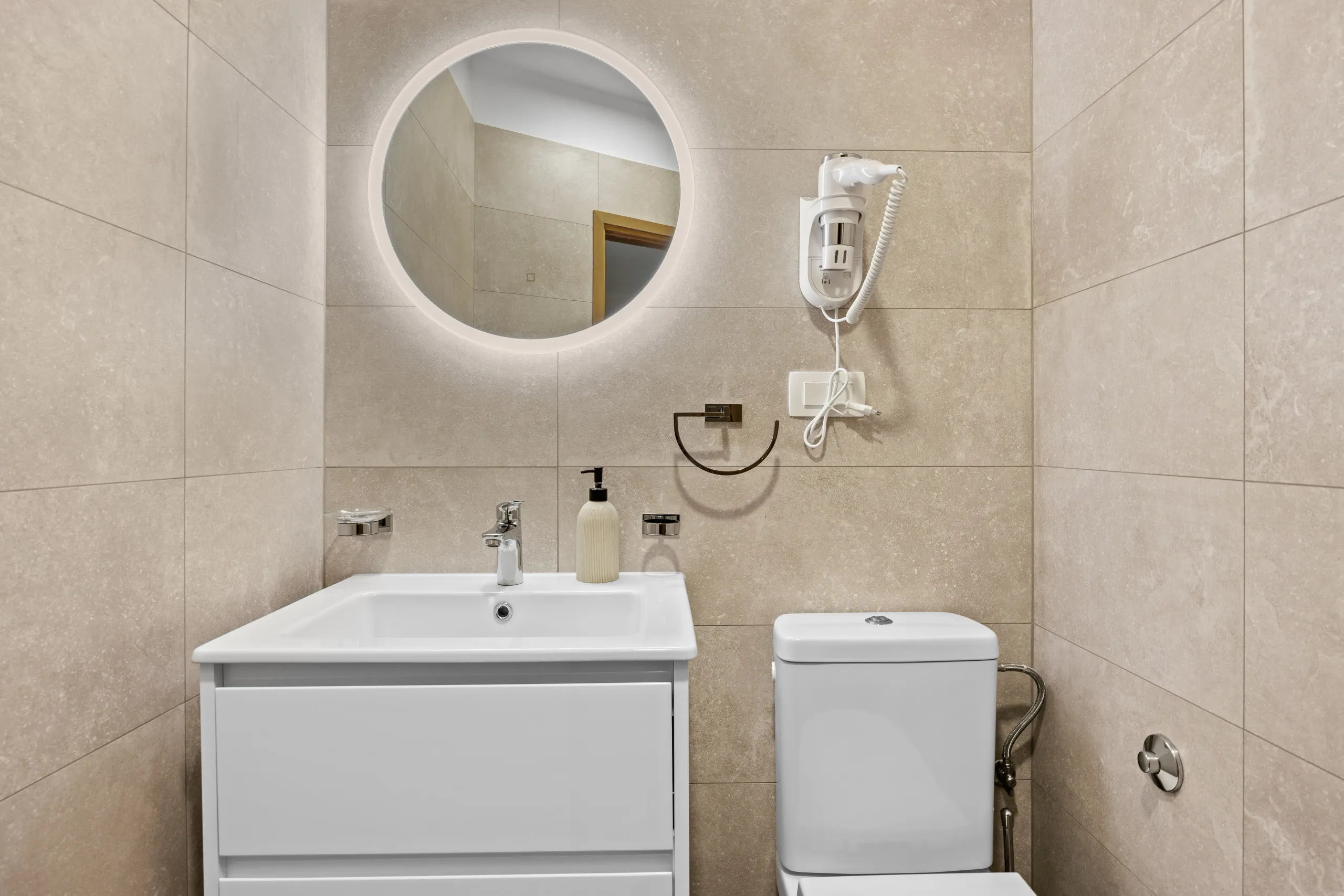 A travertine-tiled bathroom with a round backlit mirror above a white vanity